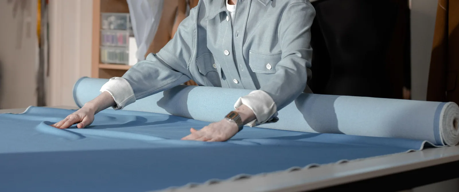 Hands smoothing blue woven fabric unrolled from a bolt across a cutting table