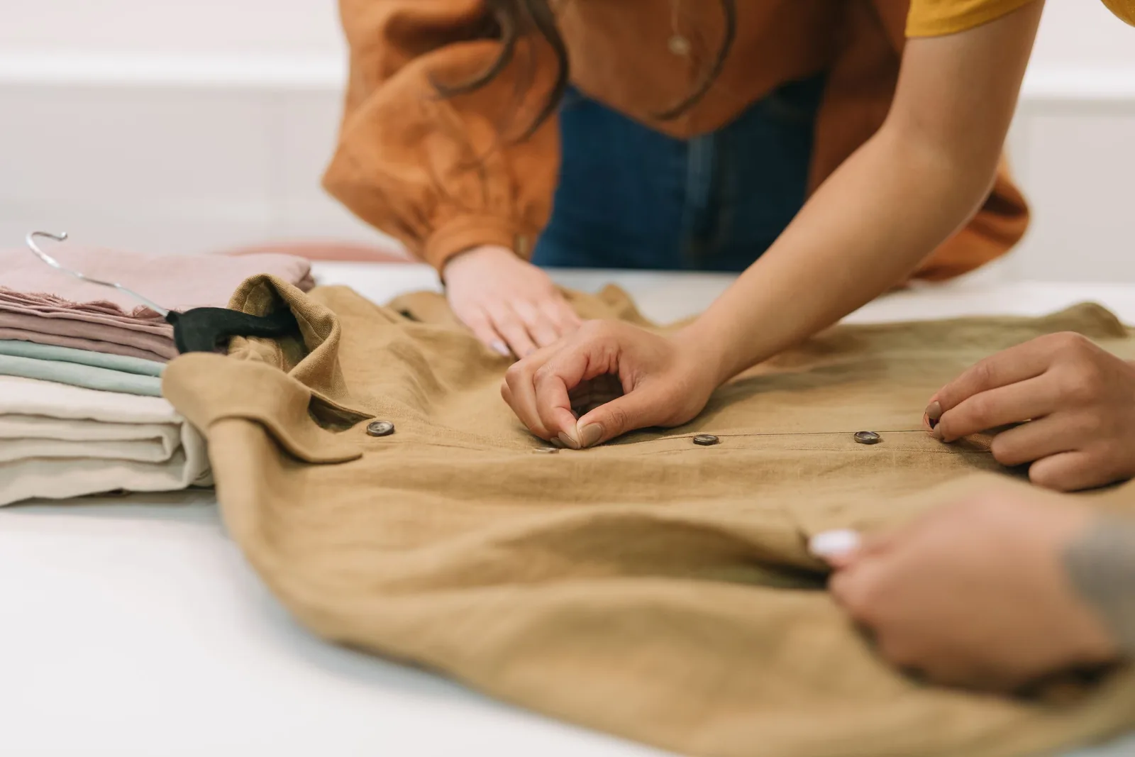 Hands checking the button placket of a linen shirt laid flat on a white table, folded garments beside it