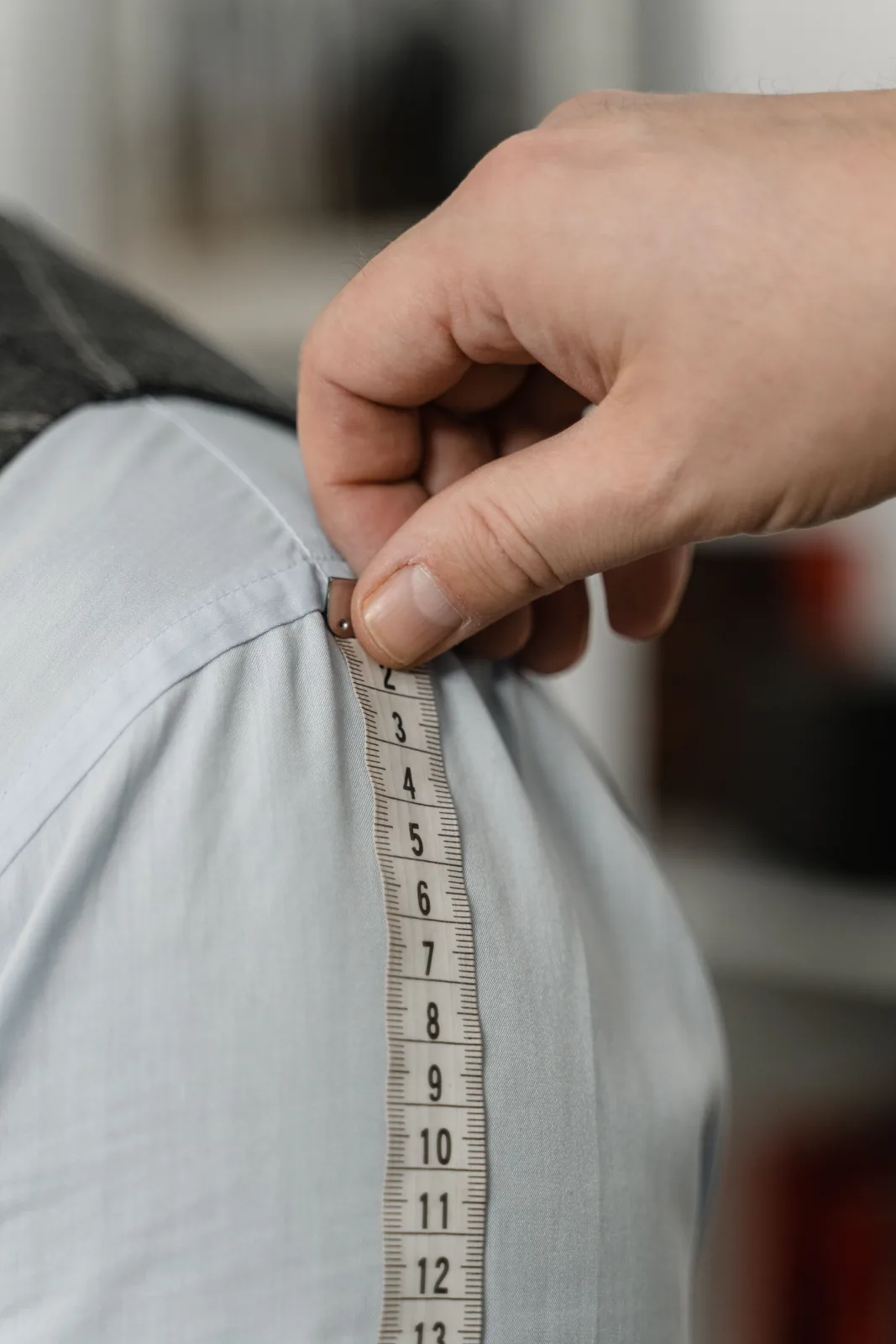 A hand holding a tape measure at the shoulder seam of a pale blue woven shirt, measuring down the sleeve