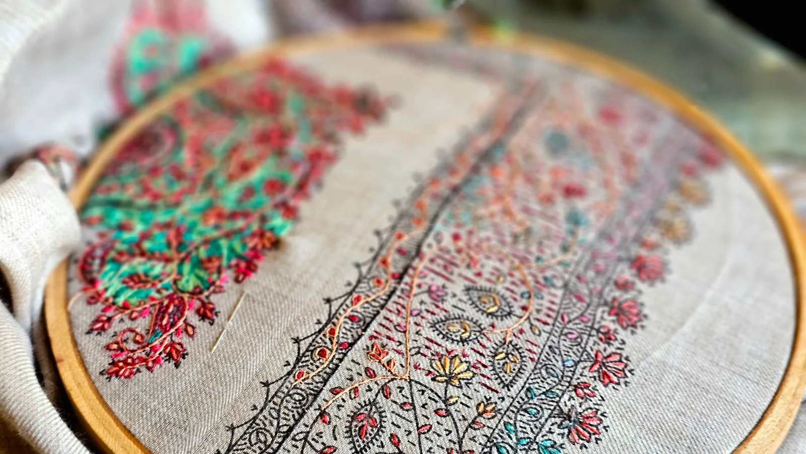 Hand embroidery in progress in a wooden hoop: dense coloured floral stitching next to a traced outline still being filled, a threaded needle resting on the cloth
