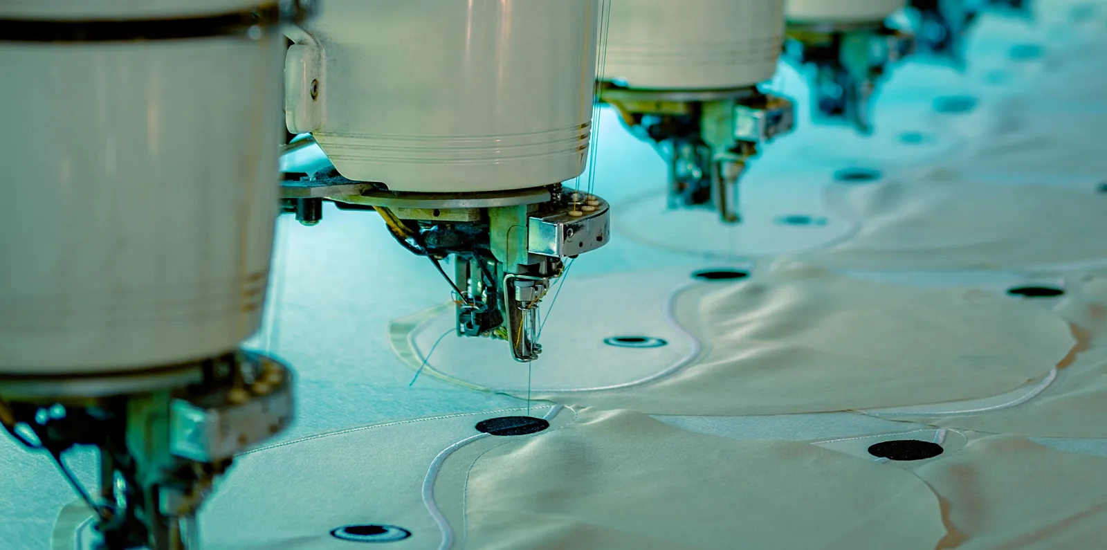 The embroidery heads of a multi-head machine in a row, stitching outlines onto cream fabric panels laid along the machine bed