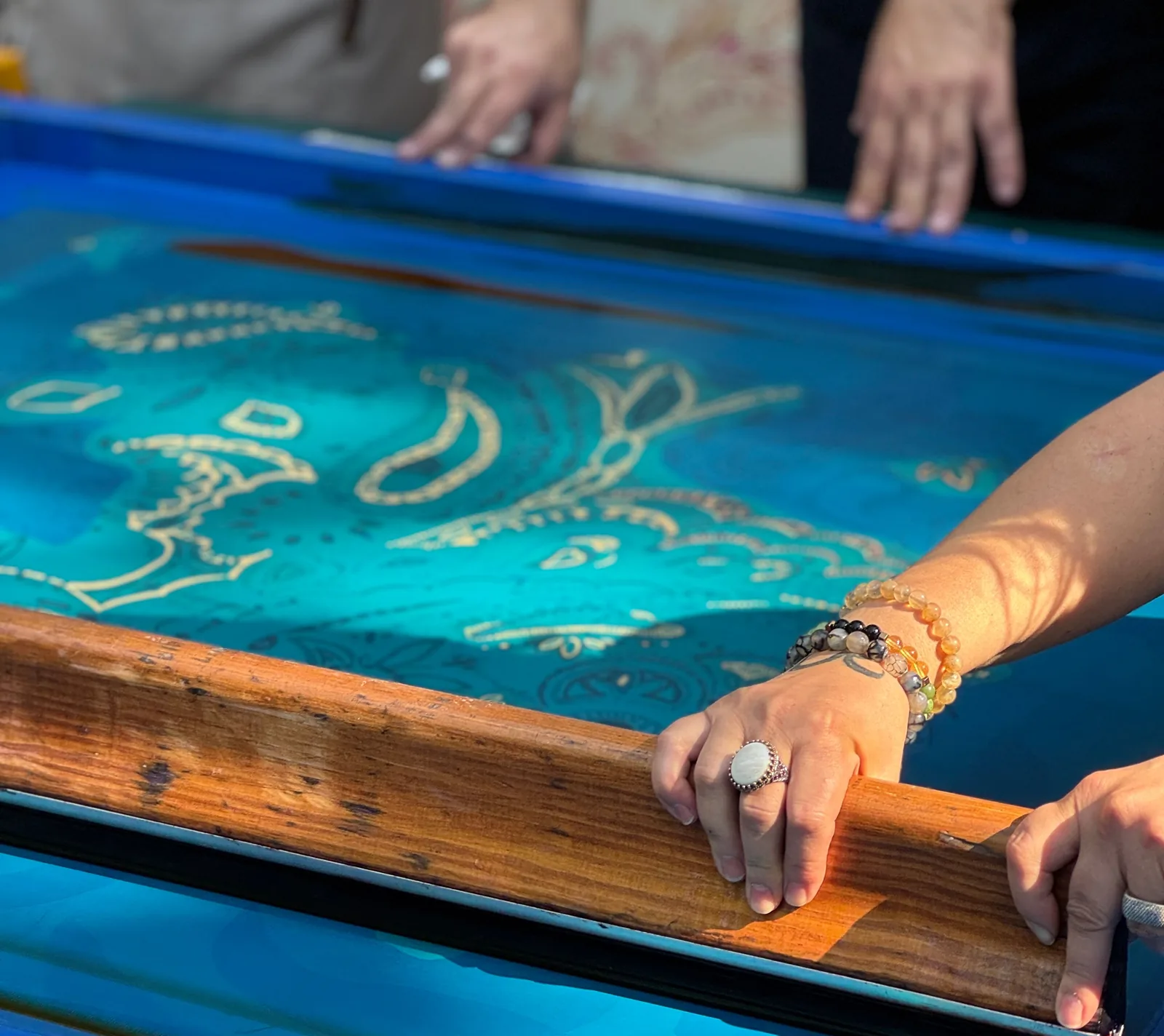 Hands hold a wooden squeegee across a blue screen-printing screen carrying a paisley design, while two more hands steady the far edge of the frame