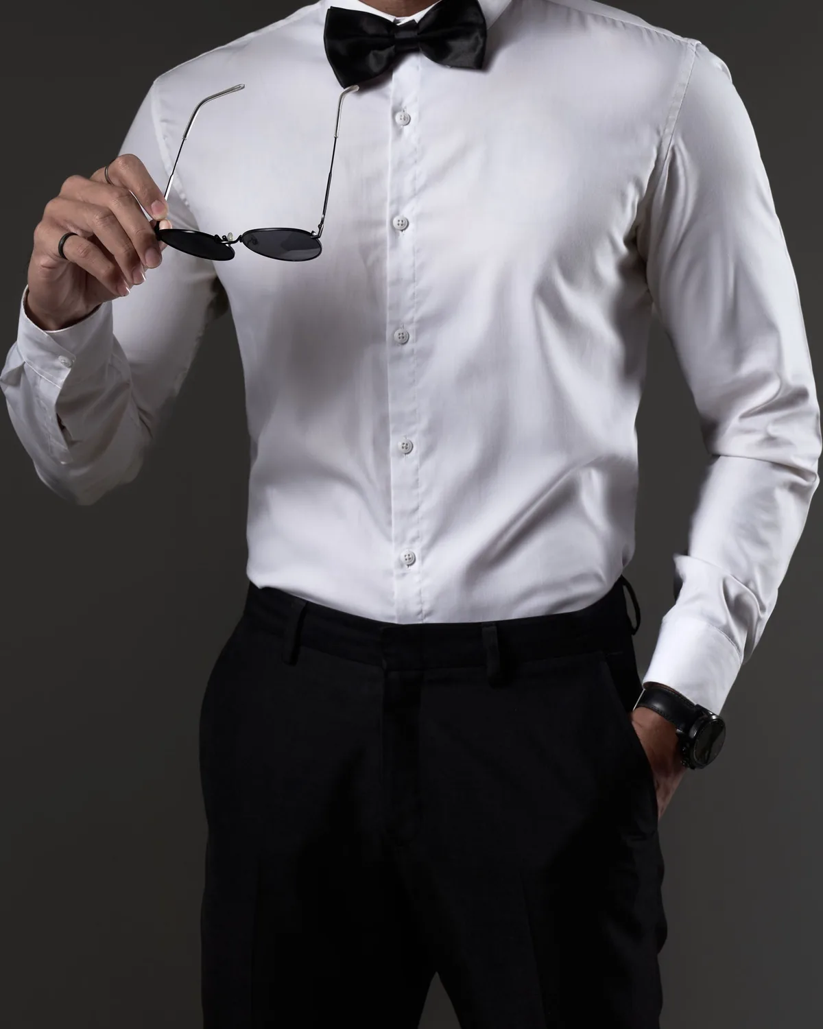 White long-sleeve dress shirt worn with a black satin bow tie, shown on a model from the neck down against a charcoal studio backdrop