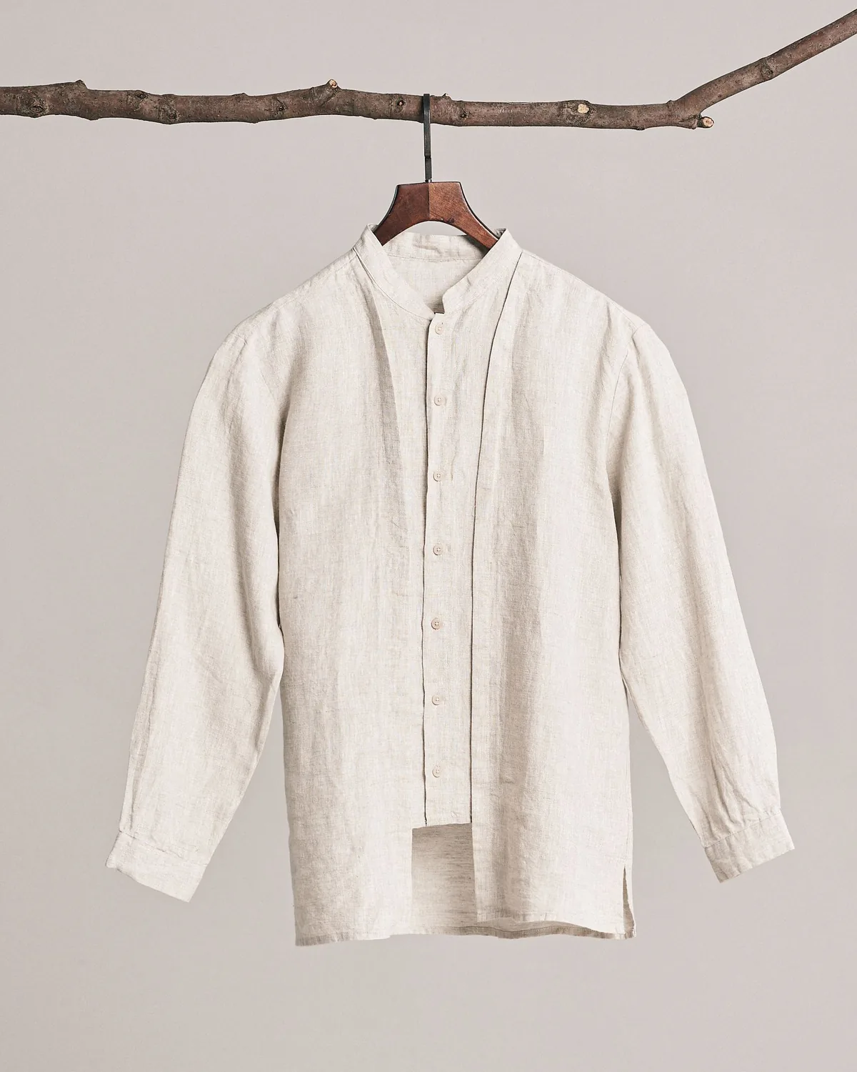 Natural oatmeal long-sleeve linen shirt with band collar and pleated front panel on a wooden hanger, slubby weave visible