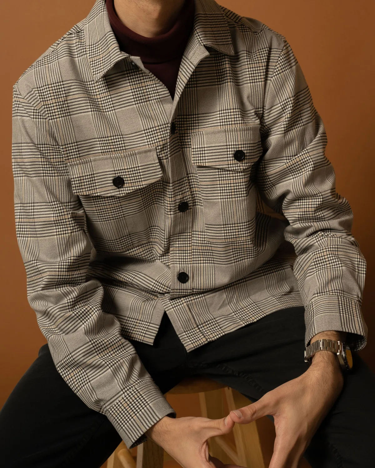 Grey glen-check overshirt with a point collar, two buttoned chest flap pockets and black buttons, worn over a burgundy roll-neck, seated and cropped below the chin on a tan studio background