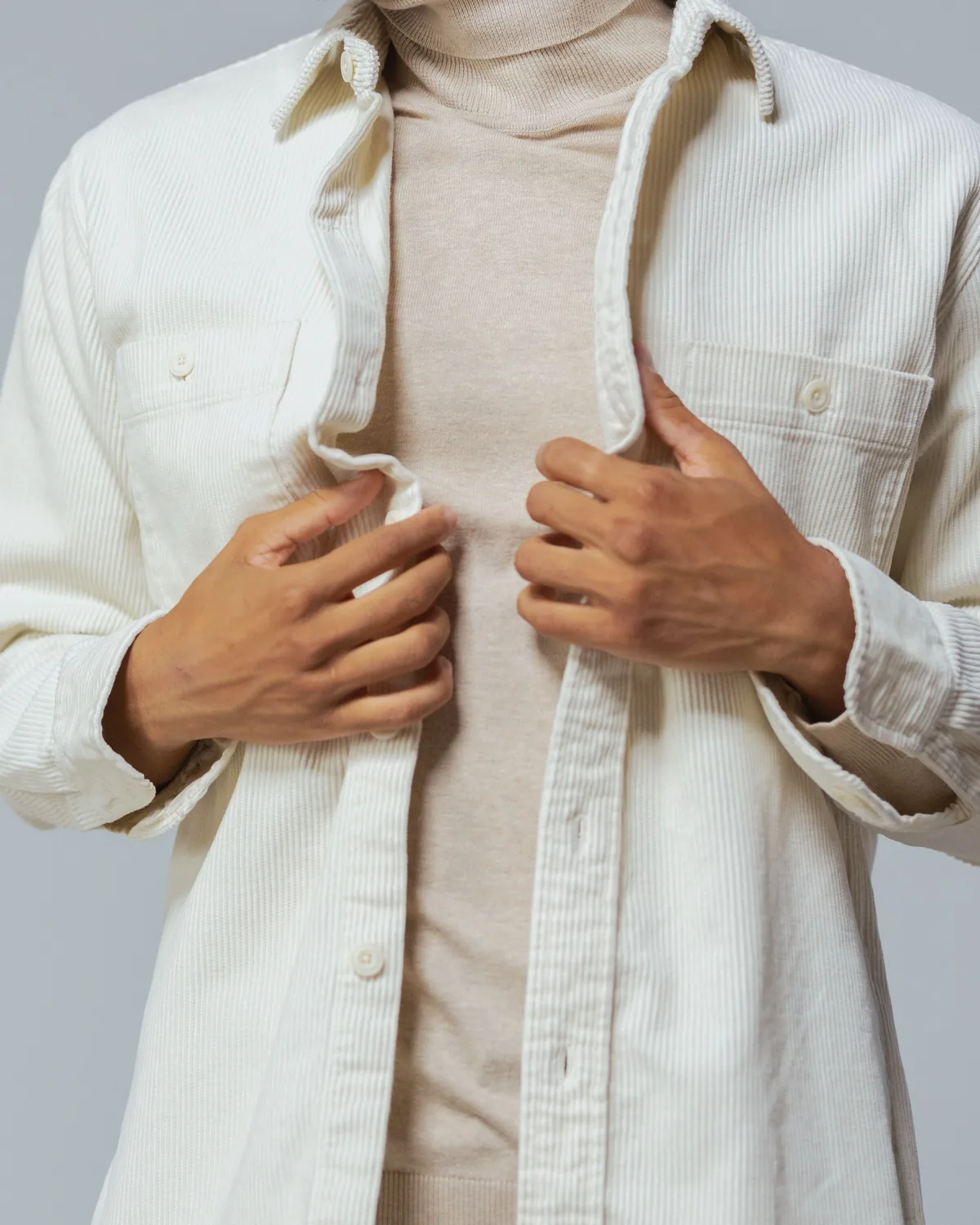 Off-white corduroy overshirt with a point collar, two buttoned chest pockets and pale buttons, worn open over a beige roll-neck, cropped below the chin on a grey studio background