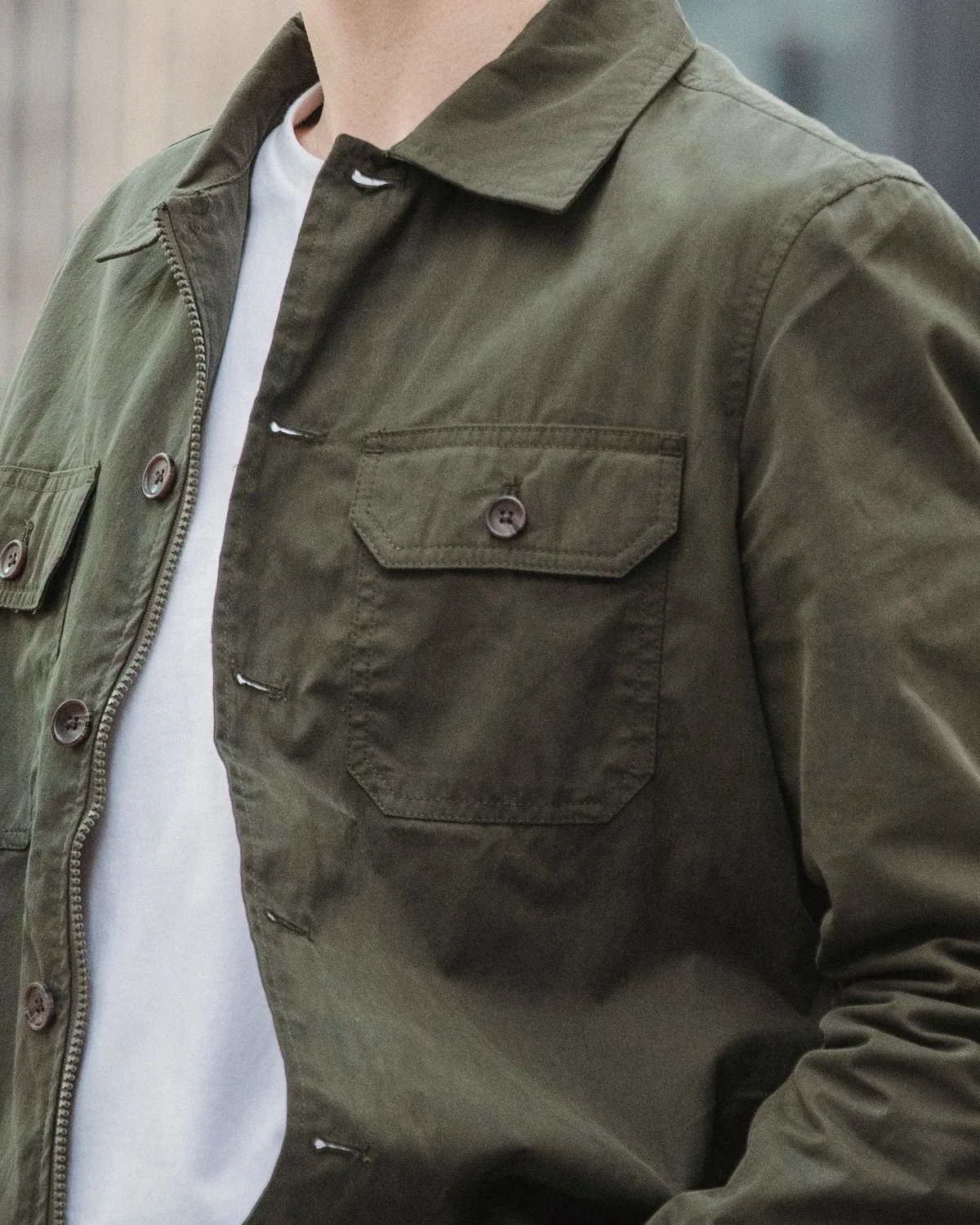 Olive cotton-twill utility overshirt with a point collar, buttoned chest flap pockets and a zip beside the button front, worn open over a white T-shirt, close crop of the chest and collar against a blurred grey street