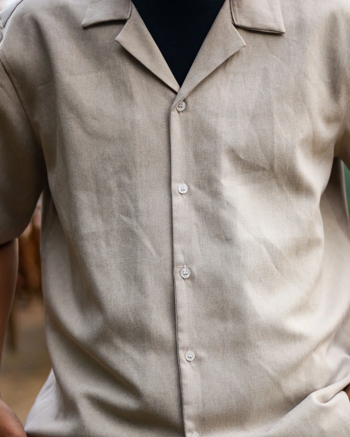 Beige short-sleeve twill shirt with an open camp collar and white four-hole buttons, worn over a black T-shirt, cropped below the chin