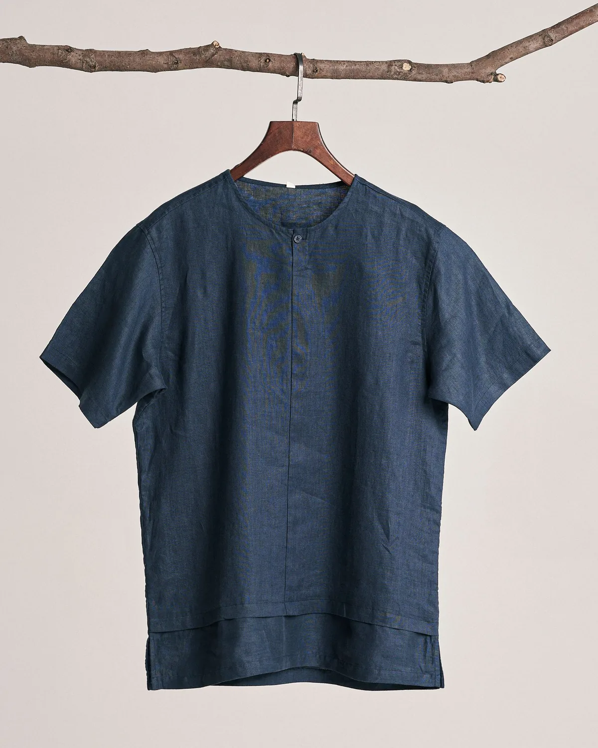 Navy short-sleeve linen-look popover shirt with a round band neck and a one-button placket, on a wooden hanger hung from a branch