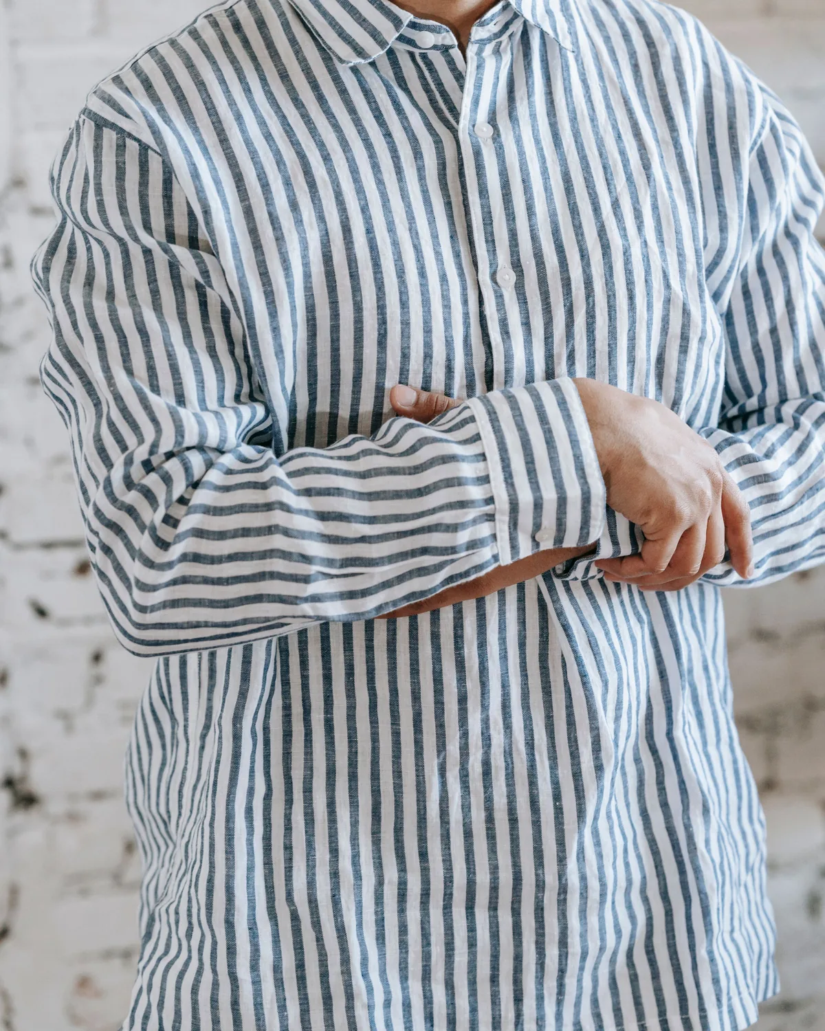 Blue and white wide-stripe popover shirt in a slubby linen-look weave, with a point collar and buttoned cuffs, worn with folded arms against a whitewashed brick wall