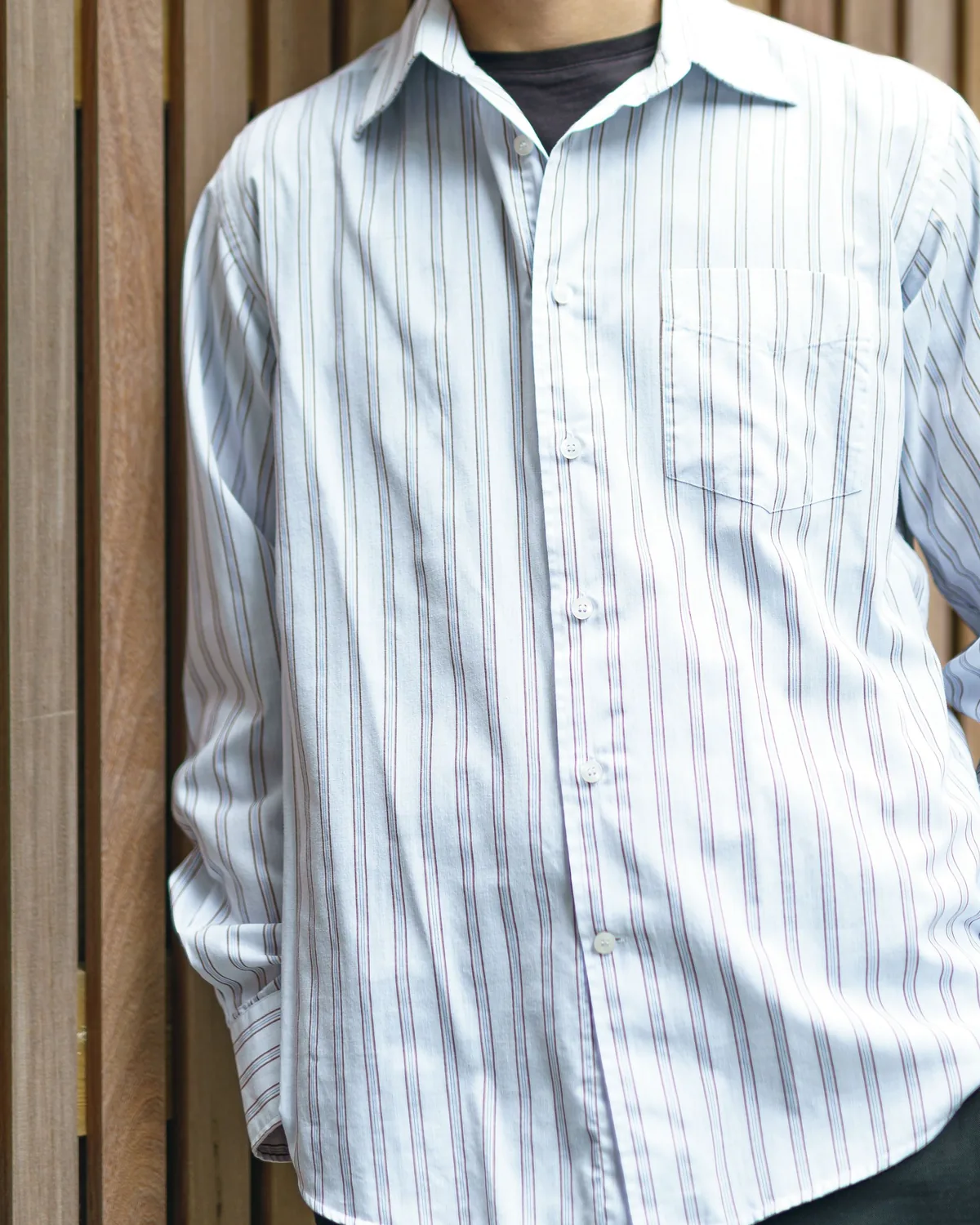 White long-sleeve shirt with fine blue and maroon pinstripes and a chest pocket, worn open at the collar against a wooden slat wall